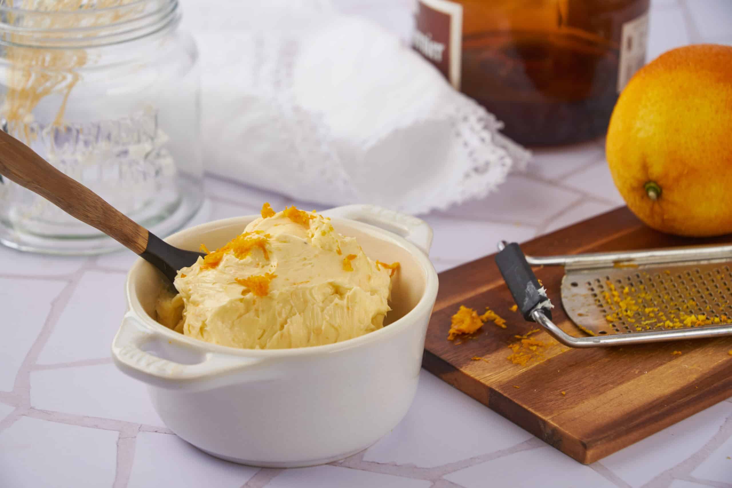 Creamy homemade vanilla ice cream in a white bowl with a wooden-handled spoon, surrounded by a grater with orange zest, fresh pear, and baking ingredients on a white tiled surface.