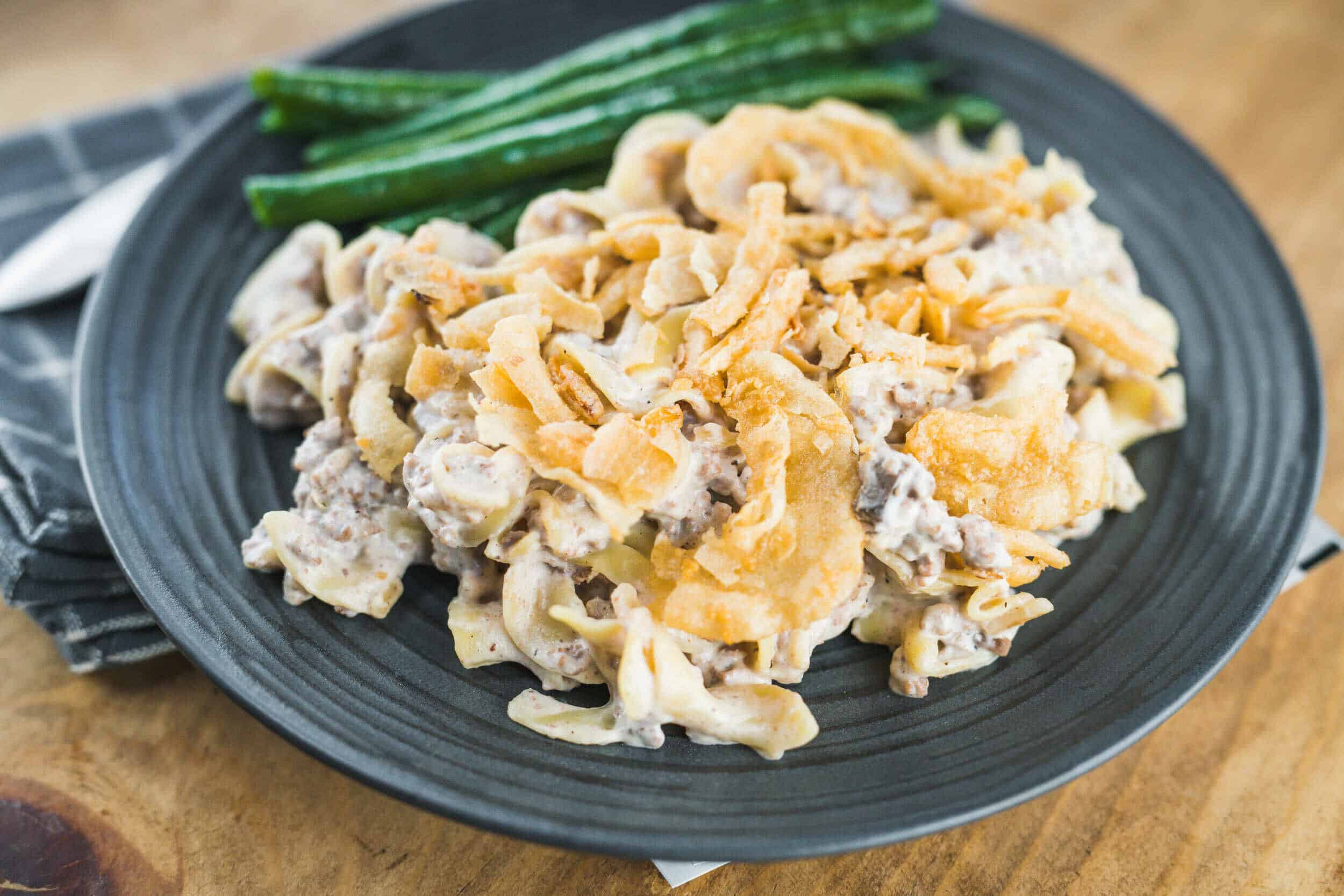 Creamy beef stroganoff with egg noodles and crispy fried onions served on a dark plate with green beans in the background.