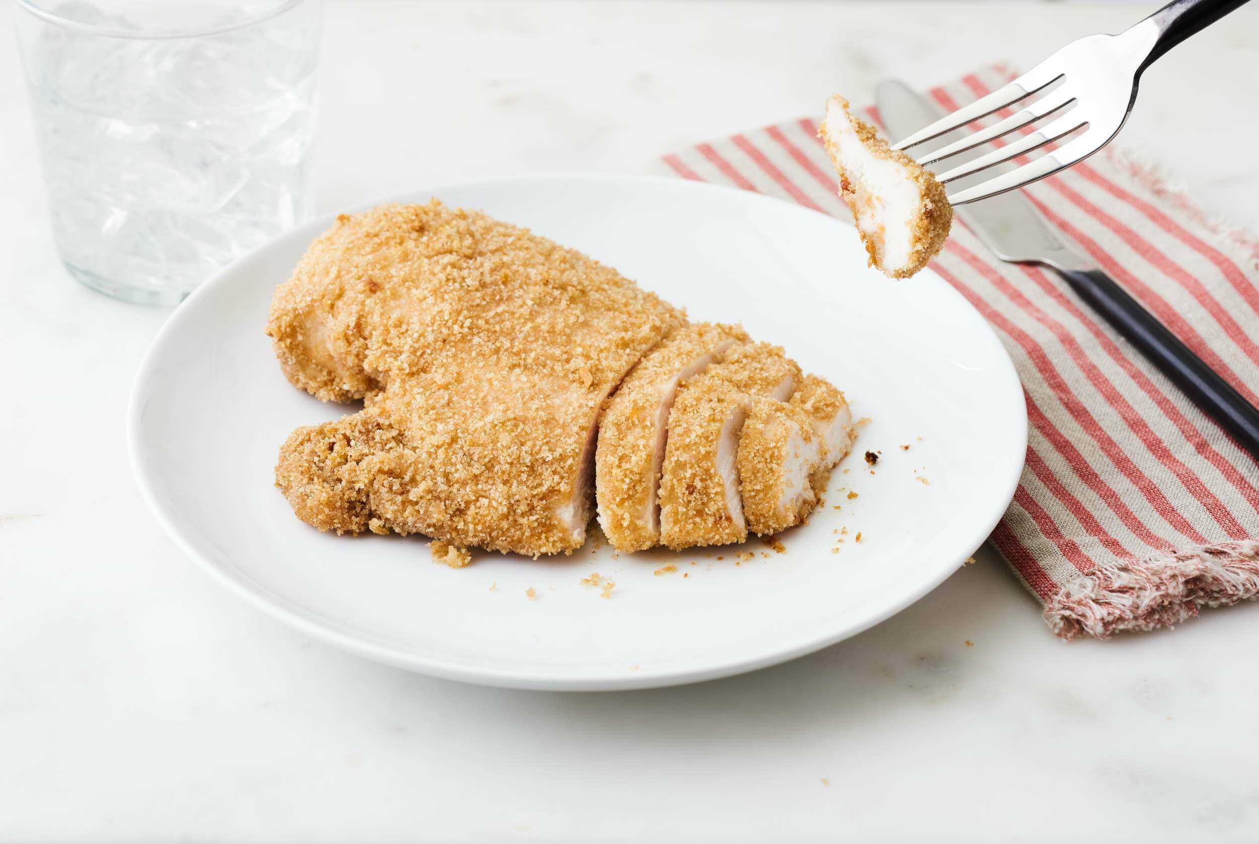 Golden fried chicken cutlet on a white plate with a glass of water in the background, served with a fork and knife on a striped cloth napkin - perfect for crispy chicken recipes and easy dinner ideas.