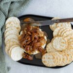 Butterscotch and pecan cheesecake topped with caramel sauce, served with round crackers on a black platter, garnished with a textured green cloth for an appealing dessert presentation.