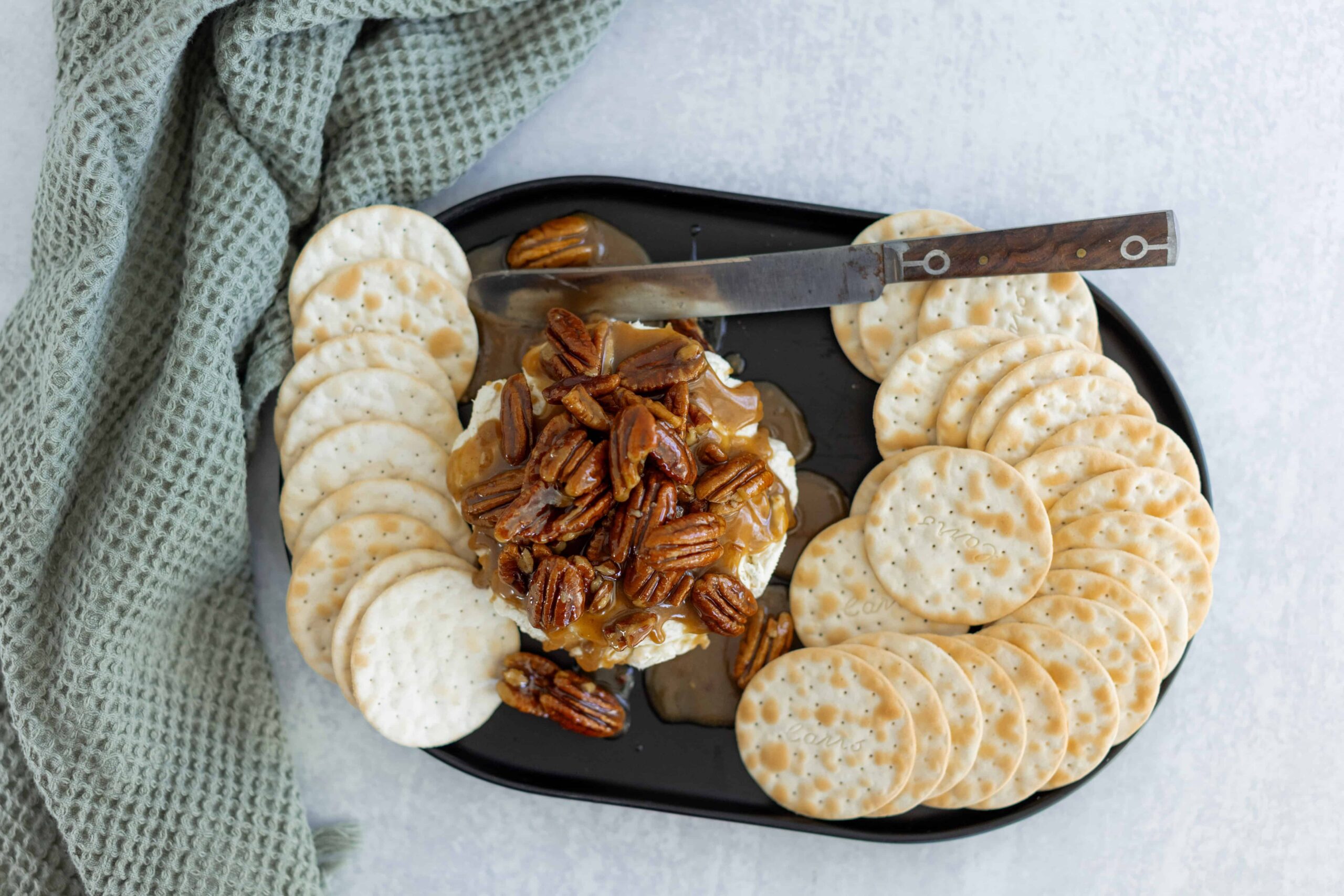 Butterscotch and pecan cheesecake topped with caramel sauce, served with round crackers on a black platter, garnished with a textured green cloth for an appealing dessert presentation.