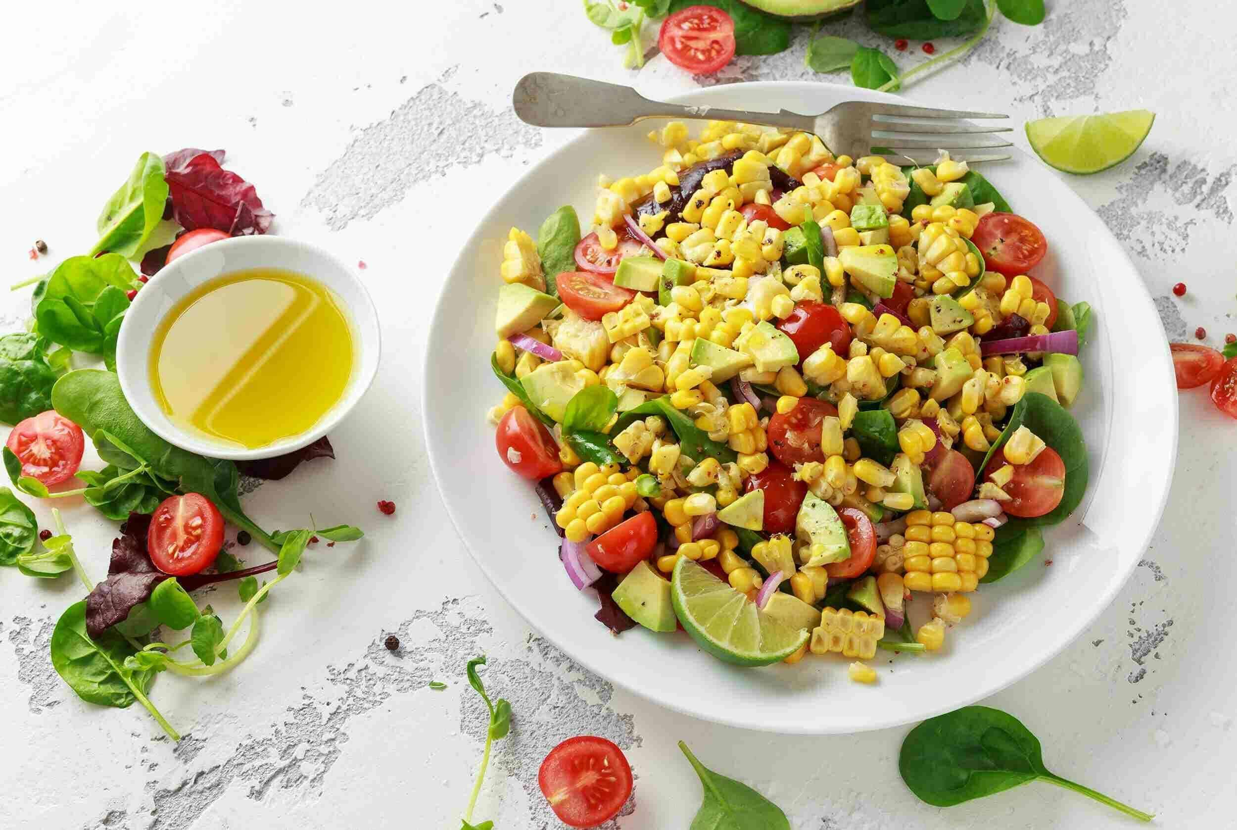 Fresh corn salad with cherry tomatoes, avocado, red onion, and lime on a white plate, served with a side of olive oil and mixed greens on a white textured surface.