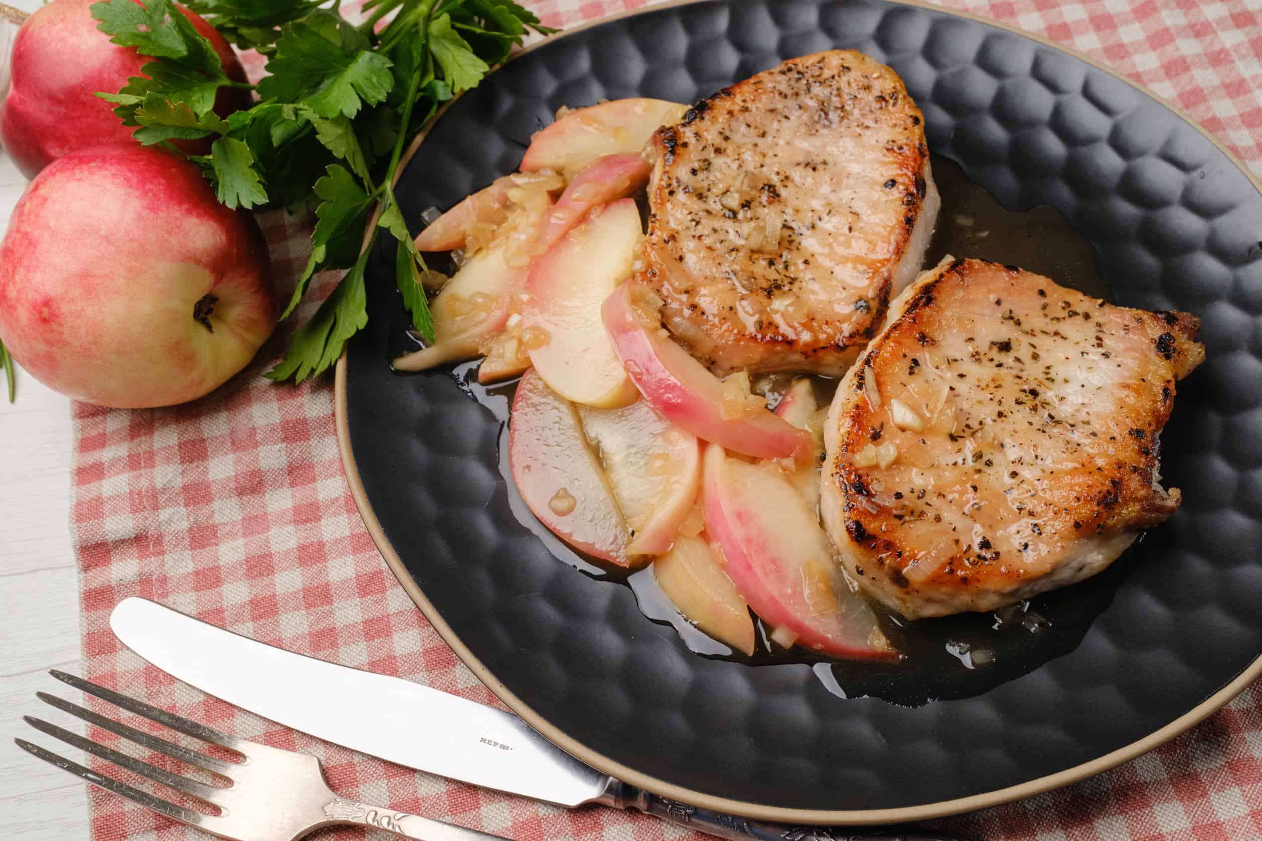 Tender grilled pork chops served with caramelized apples and onions on a black plate, fresh apples and parsley garnish, perfect for easy family dinner recipes.