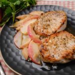 Juicy pan-seared pork chops served with sautéed apples and onions on a dark textured plate, garnished with fresh parsley, with a red apple and checkered tablecloth in the background.