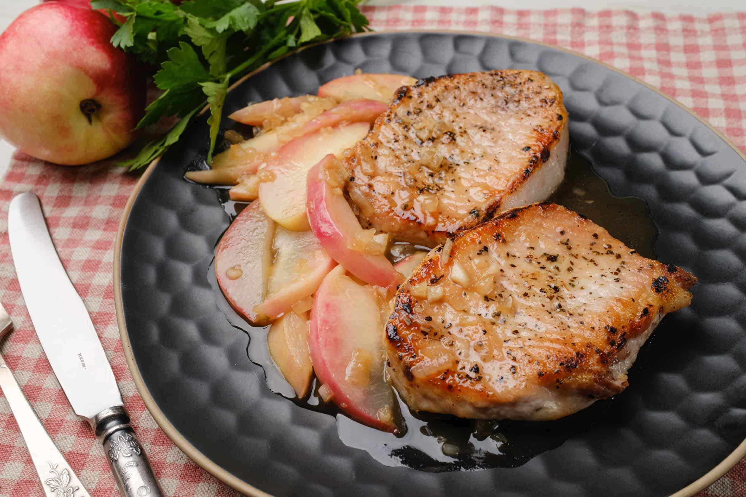 Juicy pan-seared pork chops served with sautéed apples and onions on a dark textured plate, garnished with fresh parsley, with a red apple and checkered tablecloth in the background.