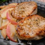 Juicy grilled pork chops with apples and caramelized onions served on a black plate. Perfect for a delicious and easy dinner recipe.