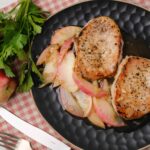 Grilled pork chops with apple onion sauté on a black plate, served with fresh herbs and apples for a delicious homemade meal.