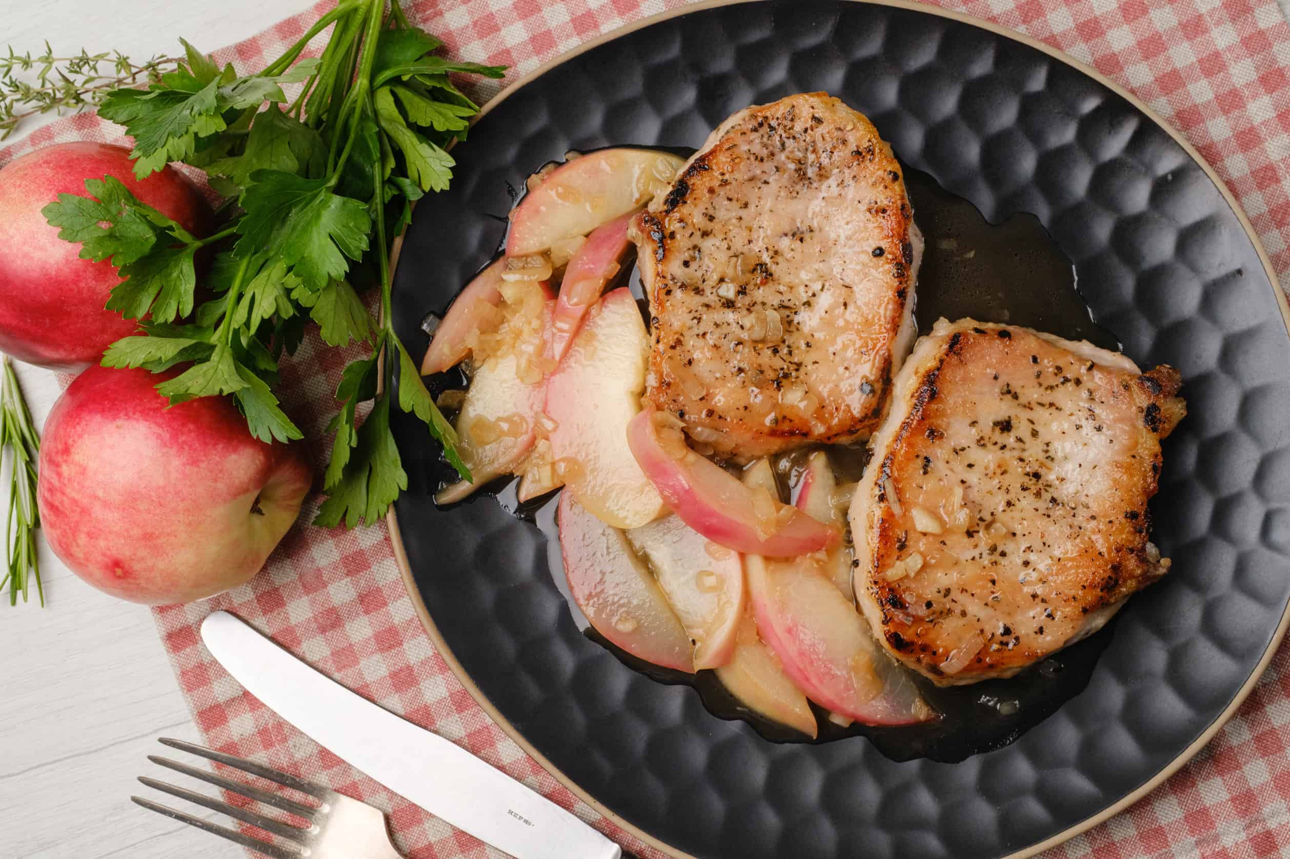 Grilled pork chops with apple onion sauté on a black plate, served with fresh herbs and apples for a delicious homemade meal.