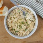 Creamy chicken alfredo pasta in a white bowl, garnished with chopped herbs, served with a side of toasted bread slices, on a wooden table with a striped cloth.