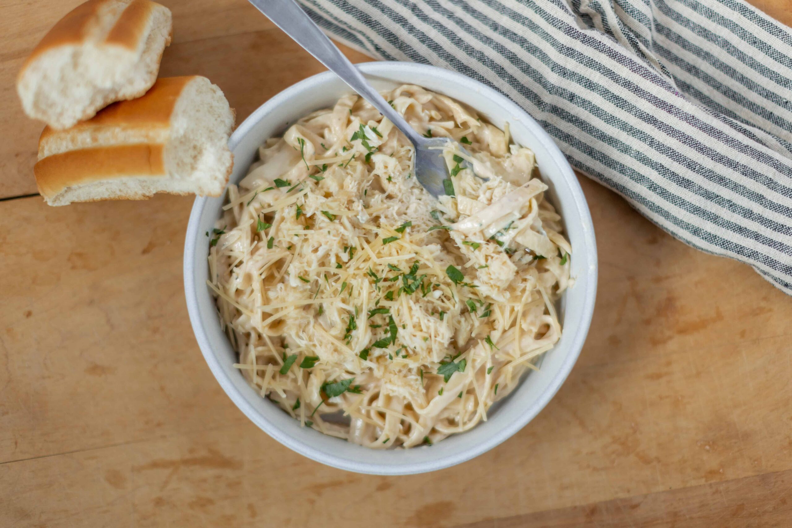 Creamy chicken alfredo pasta in a white bowl, garnished with chopped herbs, served with a side of toasted bread slices, on a wooden table with a striped cloth.