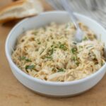 Creamy chicken pasta in a white bowl garnished with herbs on wooden surface with sliced bread in the background.