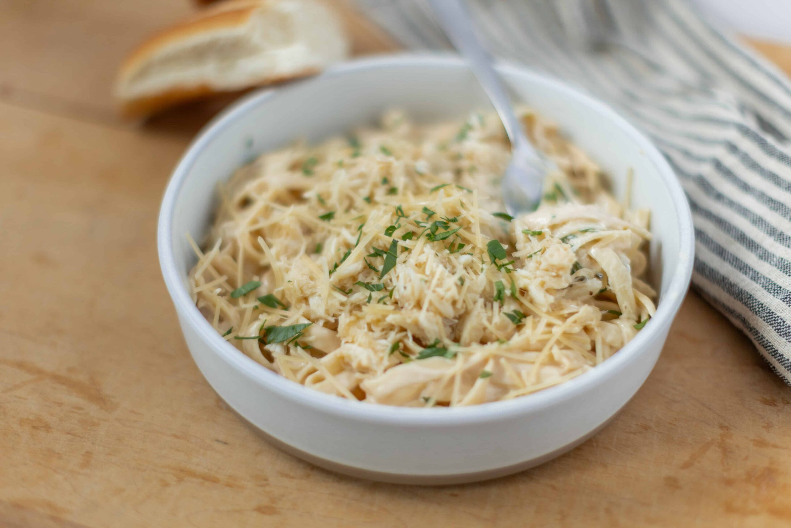 Creamy chicken pasta in a white bowl garnished with herbs on wooden surface with sliced bread in the background.