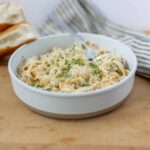 Creamy chicken Alfredo pasta topped with shredded cheese and fresh herbs in a white bowl on a wooden surface with sliced bread and a striped cloth napkin in the background.
