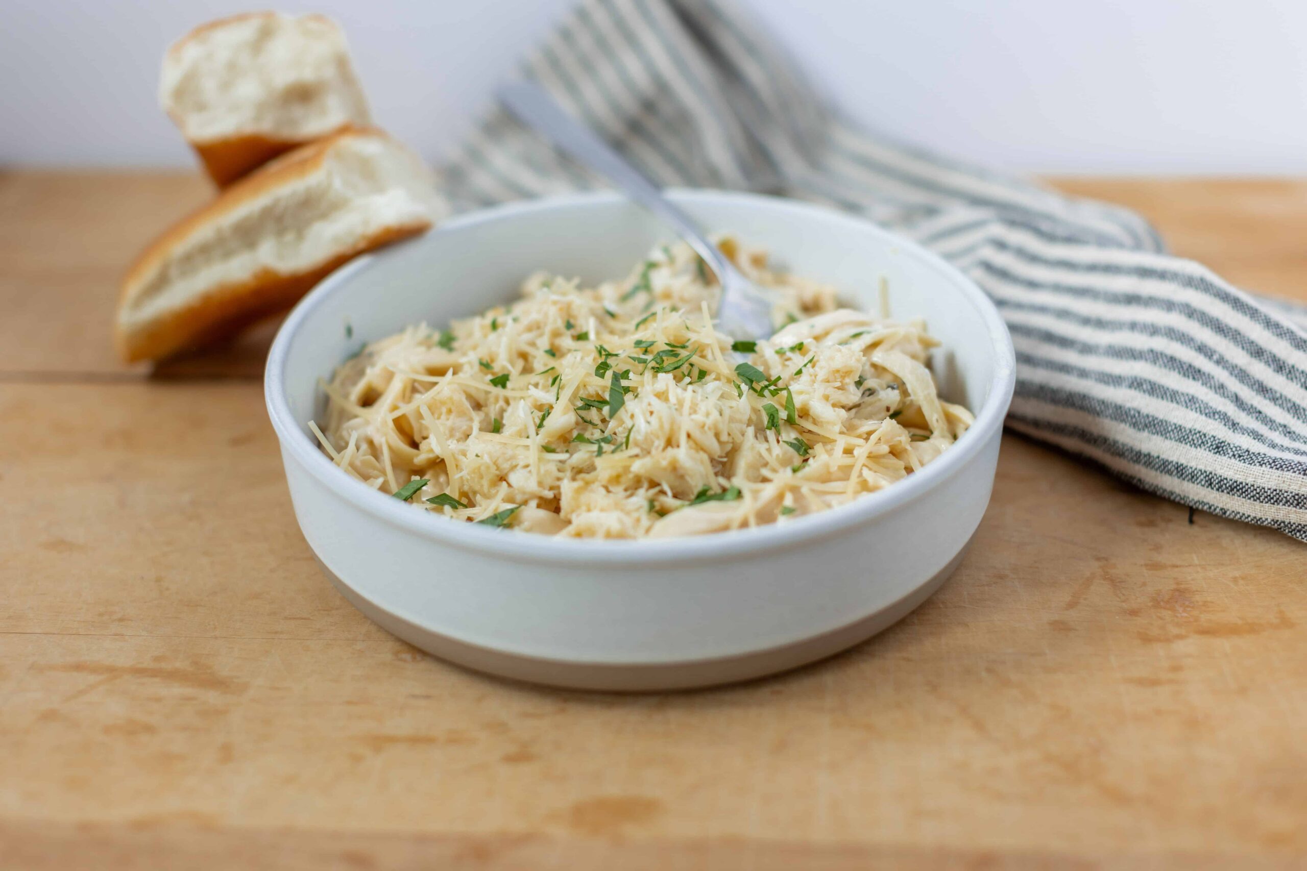 Creamy chicken Alfredo pasta topped with shredded cheese and fresh herbs in a white bowl on a wooden surface with sliced bread and a striped cloth napkin in the background.