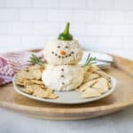 Creamy cheese ball snowman with carrot nose, green pepper hat, black pepper eyes, and rosemary arms on a wooden tray, decorated for holiday festive cheese platter, served with crackers and white background.