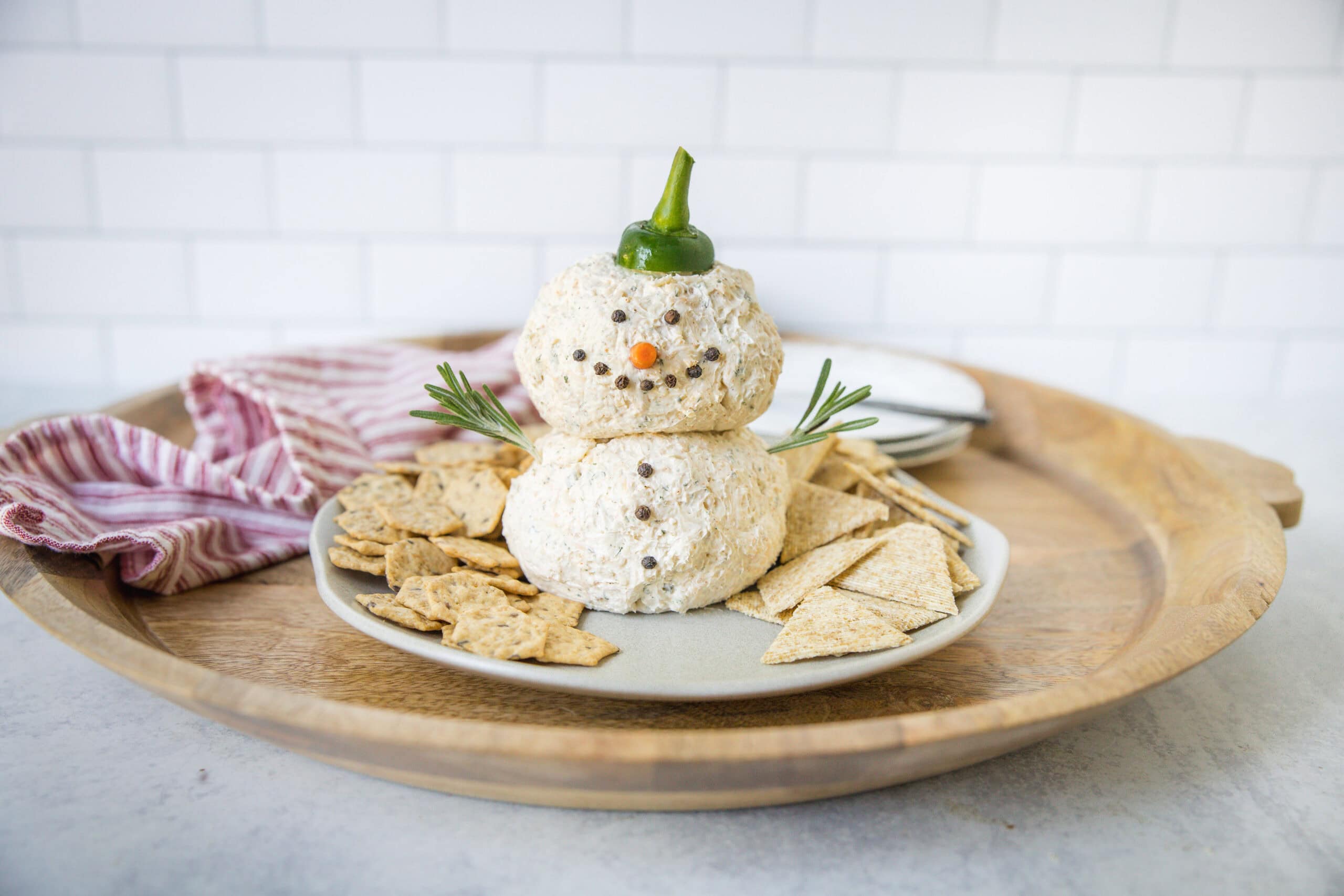 Creamy cheese ball snowman with carrot nose, green pepper hat, black pepper eyes, and rosemary arms on a wooden tray, decorated for holiday festive cheese platter, served with crackers and white background.