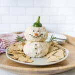 Cream cheese snowman appetizer made with cream cheese, pecans, and vegetables, decorated as a snowman for holiday party serving ideas, with crackers and herbs on a wooden tray.