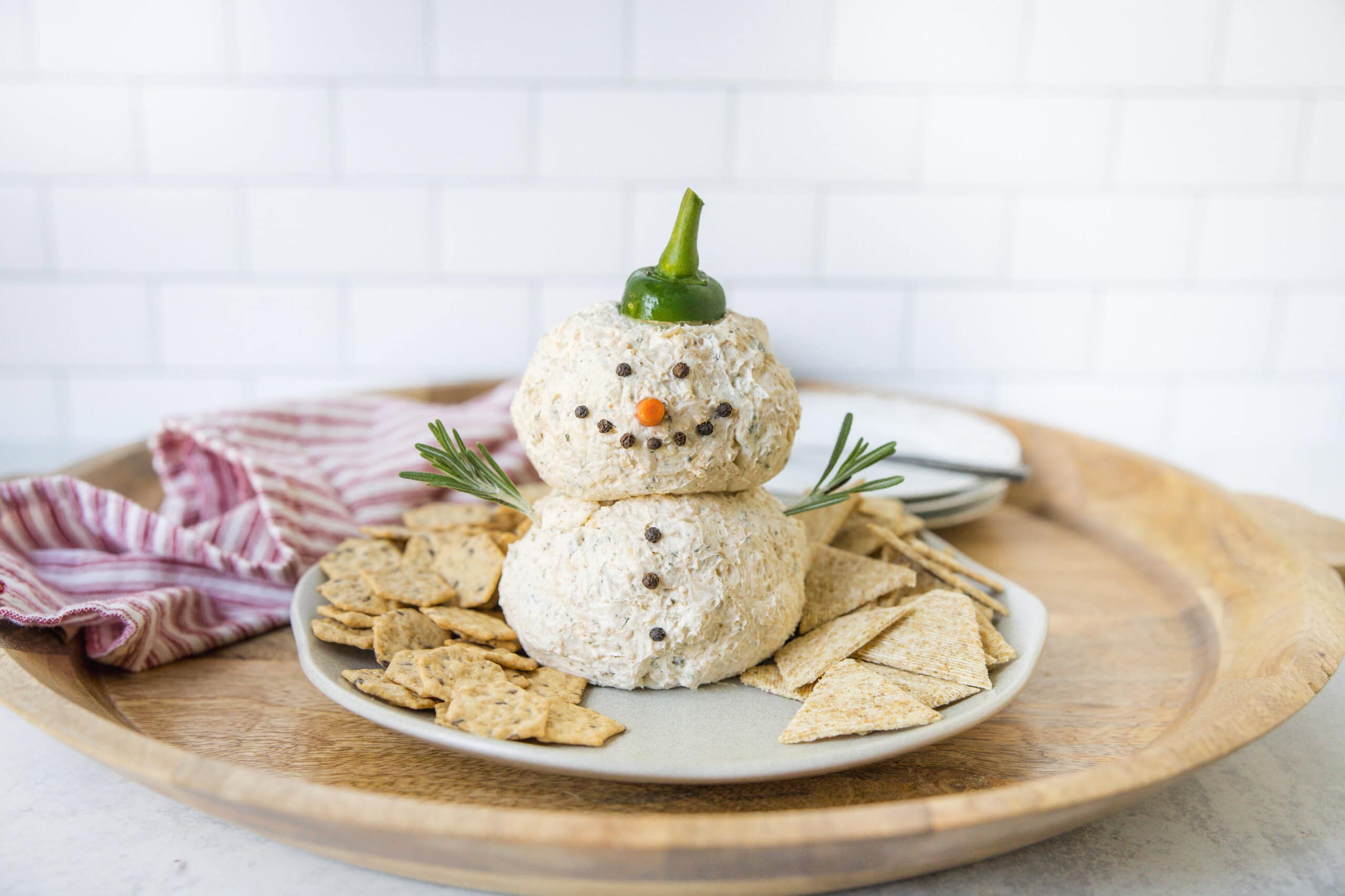 Cream cheese snowman appetizer made with cream cheese, pecans, and vegetables, decorated as a snowman for holiday party serving ideas, with crackers and herbs on a wooden tray.