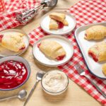 Soft homemade cherry turnovers on a baking sheet with bowls of cherry jam, whipped cream, and a fork, served on a checkered red and white tablecloth - delicious dessert recipe from Hey Recipe.