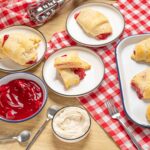 Flaky cherry turnovers with golden pastry crust served on white plates with cherry jam and whipped cream on a rustic wooden table; perfect homemade dessert.