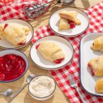 Flaky cherry-filled Danish pastries fresh out of the oven served on white plates on a wooden table with a red-checkered cloth.