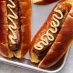 Golden-brown hot dogs in soft buns topped with mustard, served on a silver tray with mustard and relish condiments nearby.