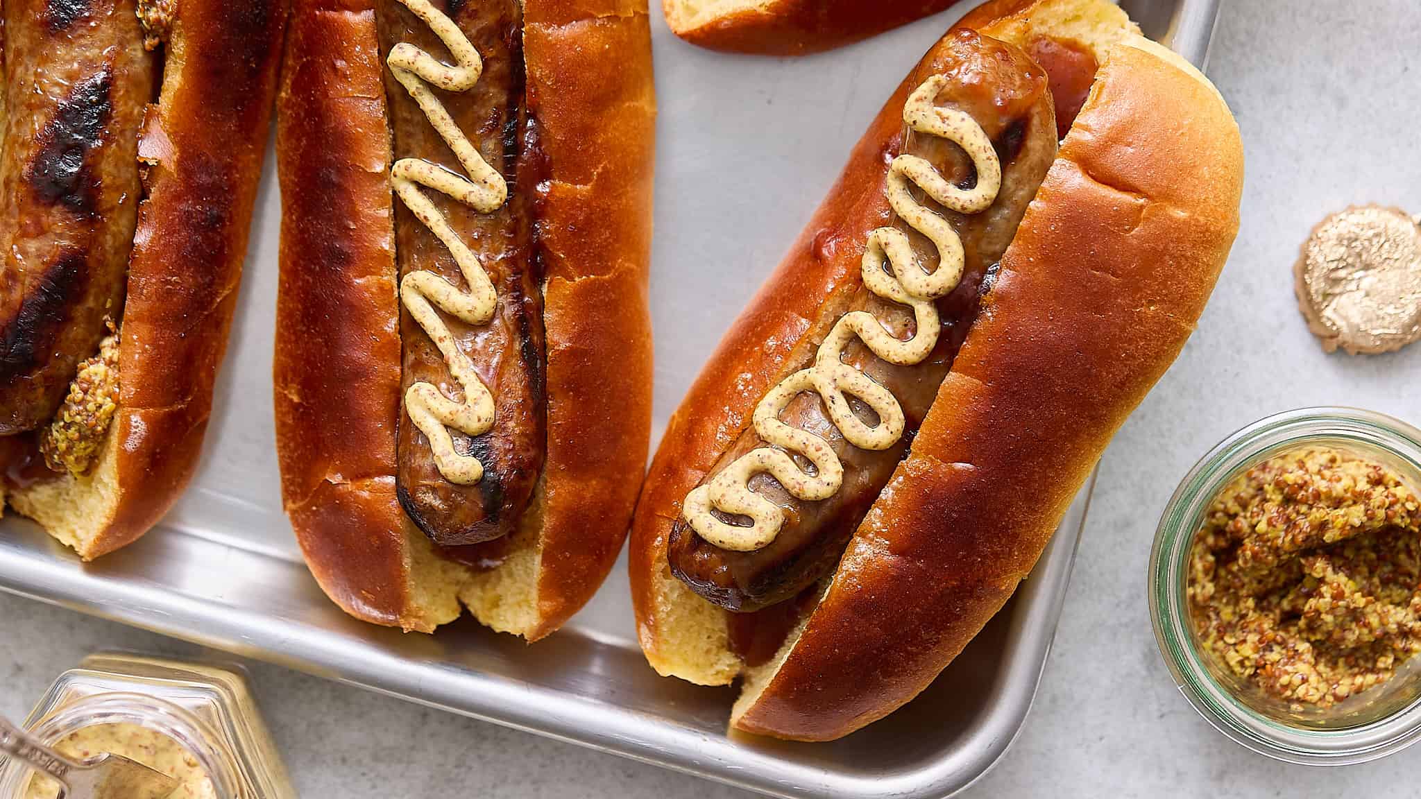 Golden-brown hot dogs in soft buns topped with mustard, served on a silver tray with mustard and relish condiments nearby.