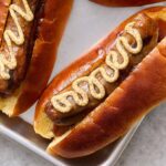 Juicy hot dogs in soft buns topped with mustard on a metal tray.