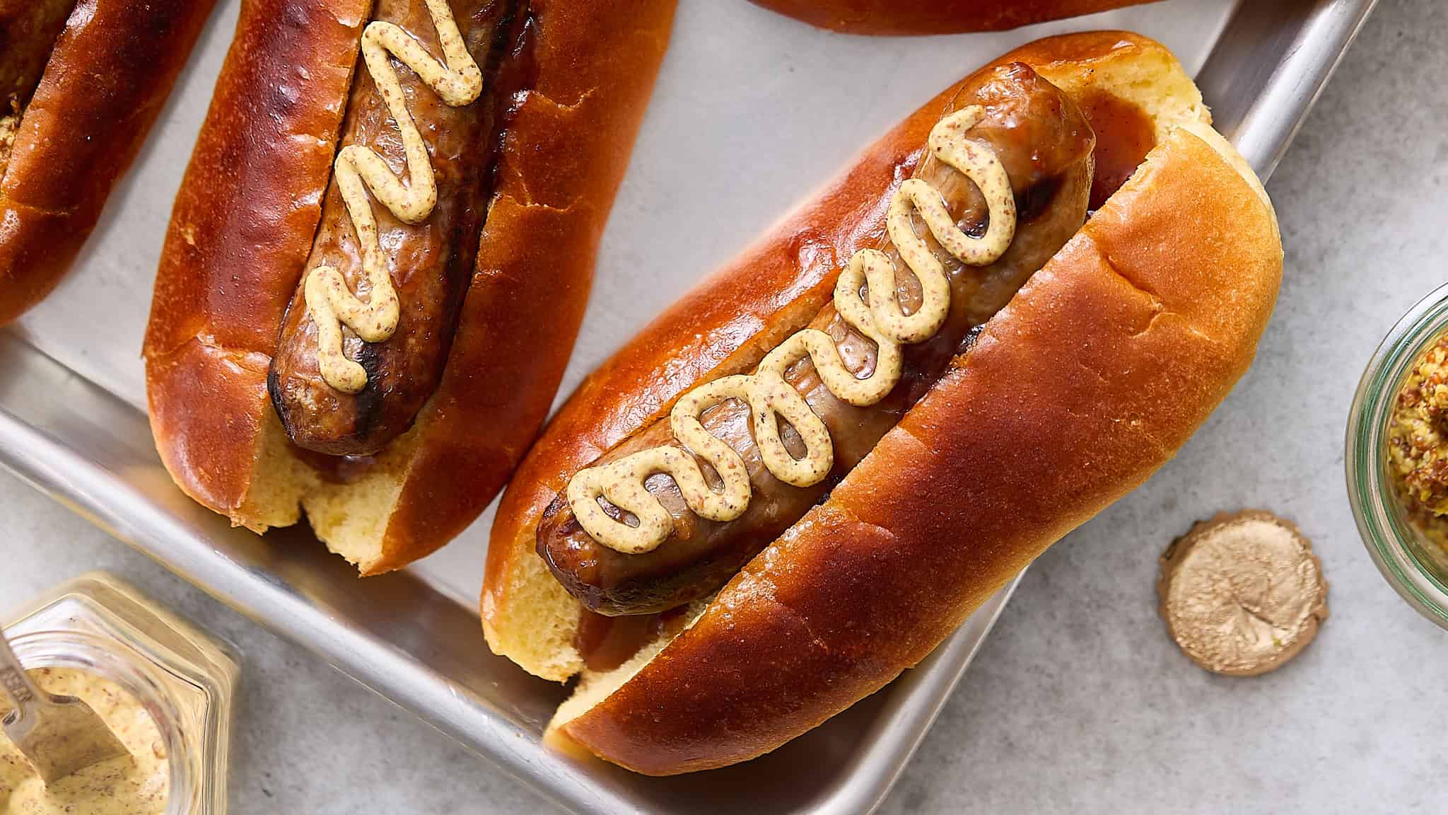 Juicy hot dogs in soft buns topped with mustard on a metal tray.