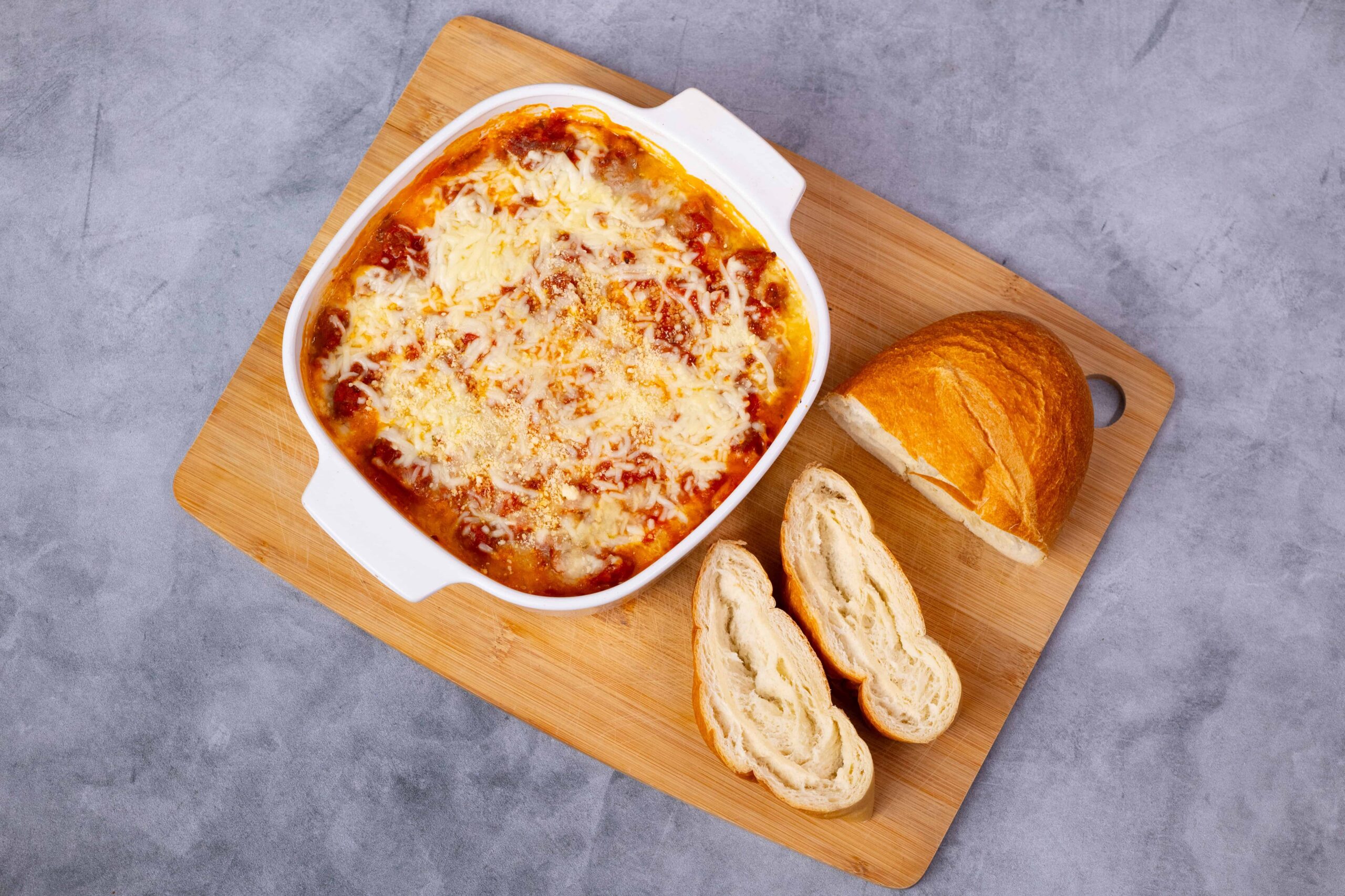 Creamy baked lasagna with melted cheese served in a white casserole dish on a wooden cutting board with fresh bread and baguette slices.