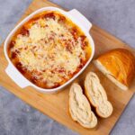 Baked cheese dip with melted cheese, tomato sauce, and breadcrumbs served in a white ceramic baking dish, accompanied by a fresh baguette bread on a wooden cutting board.