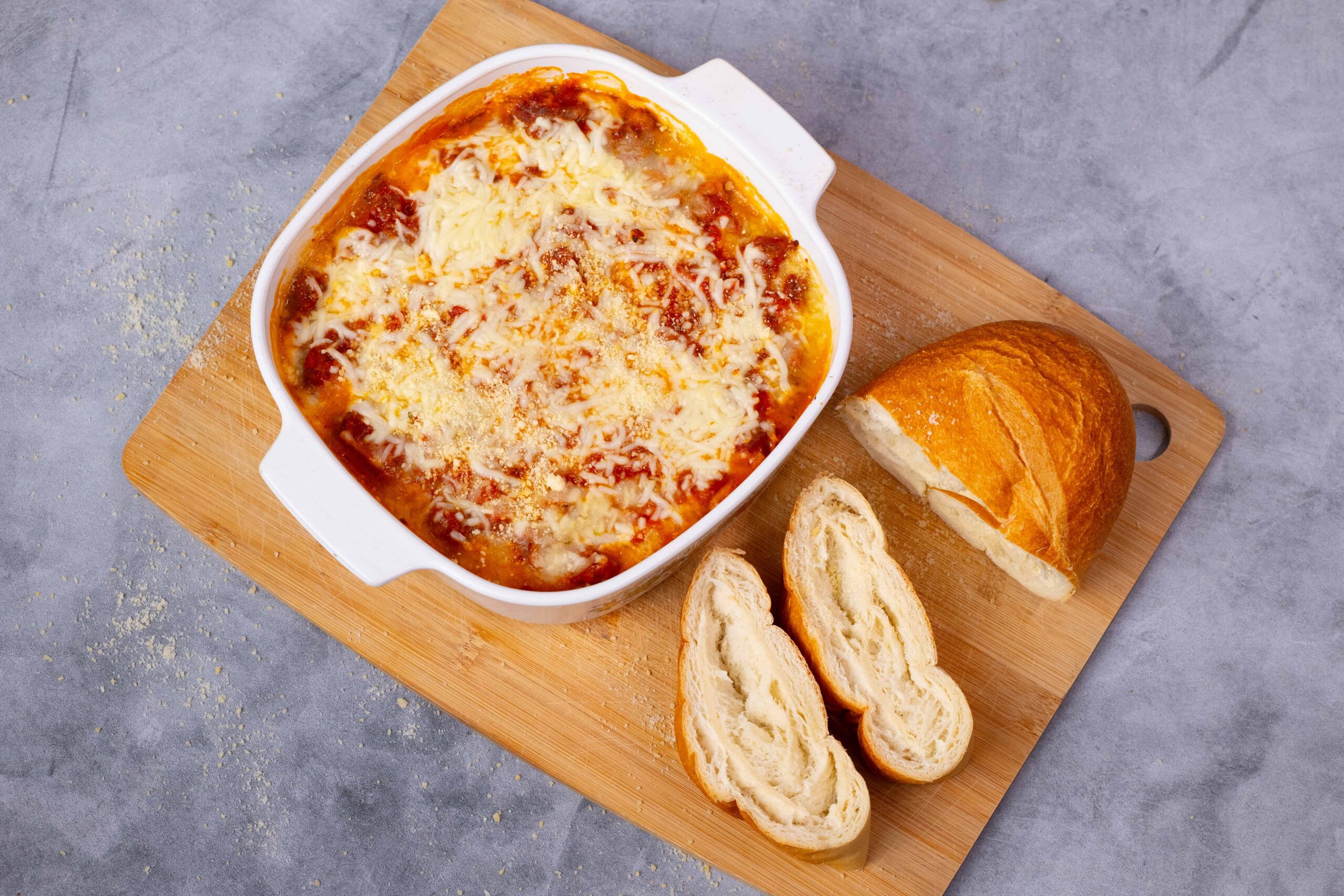 Baked cheese dip with melted cheese, tomato sauce, and breadcrumbs served in a white ceramic baking dish, accompanied by a fresh baguette bread on a wooden cutting board.