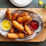 Crispy fried corn dogs served with mustard and ketchup dipping sauces on a wooden tray.
