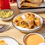 Savory Mexican beef and bean flautas served with toppings and side dishes, featuring crispy rolled tortillas filled with seasoned meat, cheese, and vegetables, on a rustic wooden table.