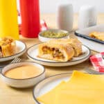 Savory cheeseburger casserole slices with beef, cheese, and breading served on a white plate surrounded by condiments, cheese slices, and side dishes on a wooden table.