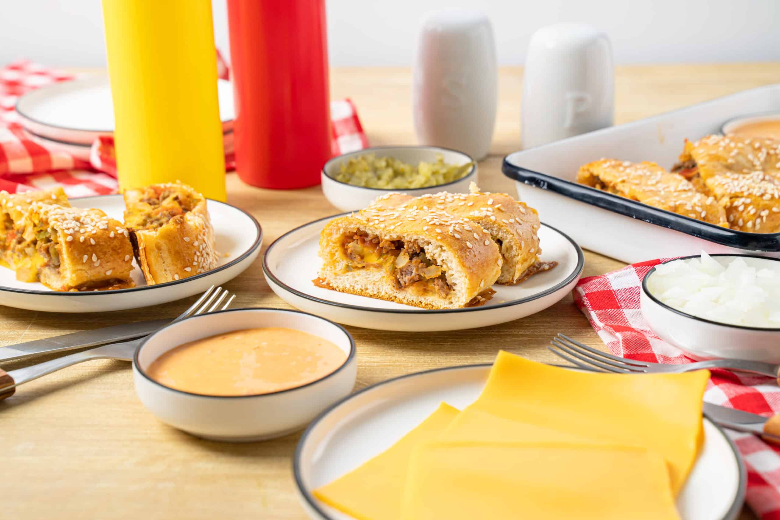 Savory cheeseburger casserole slices with beef, cheese, and breading served on a white plate surrounded by condiments, cheese slices, and side dishes on a wooden table.