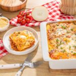 Baked lasagna with melted cheese, fresh tomato ingredients, and a side of grated Parmesan, served on a rustic wooden table with red checkered cloth and kitchen utensils.