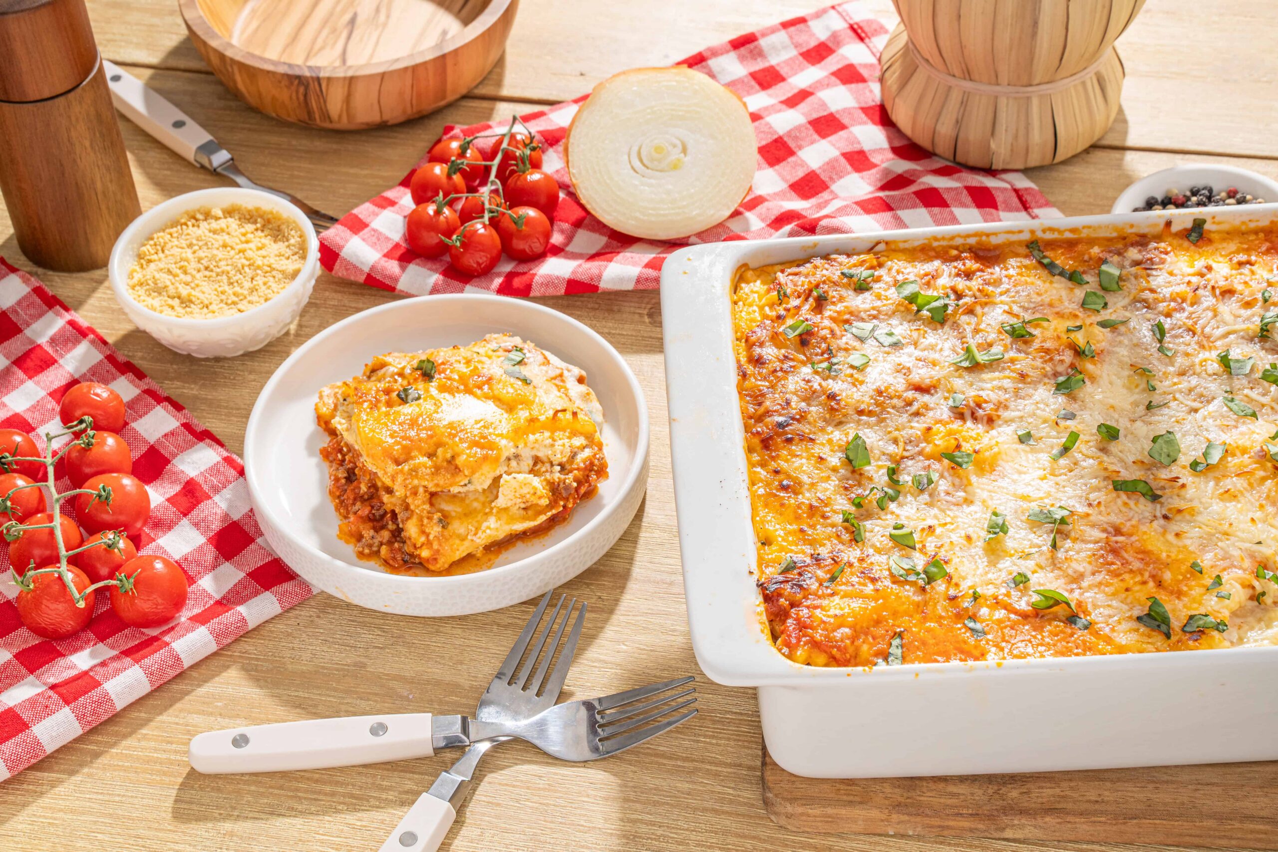 Baked lasagna with melted cheese, fresh tomato ingredients, and a side of grated Parmesan, served on a rustic wooden table with red checkered cloth and kitchen utensils.