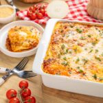 Cheesy baked lasagna with fresh herbs in a white casserole dish served with a slice on a white plate, surrounded by cherry tomatoes and cooking ingredients on a rustic wooden table.
