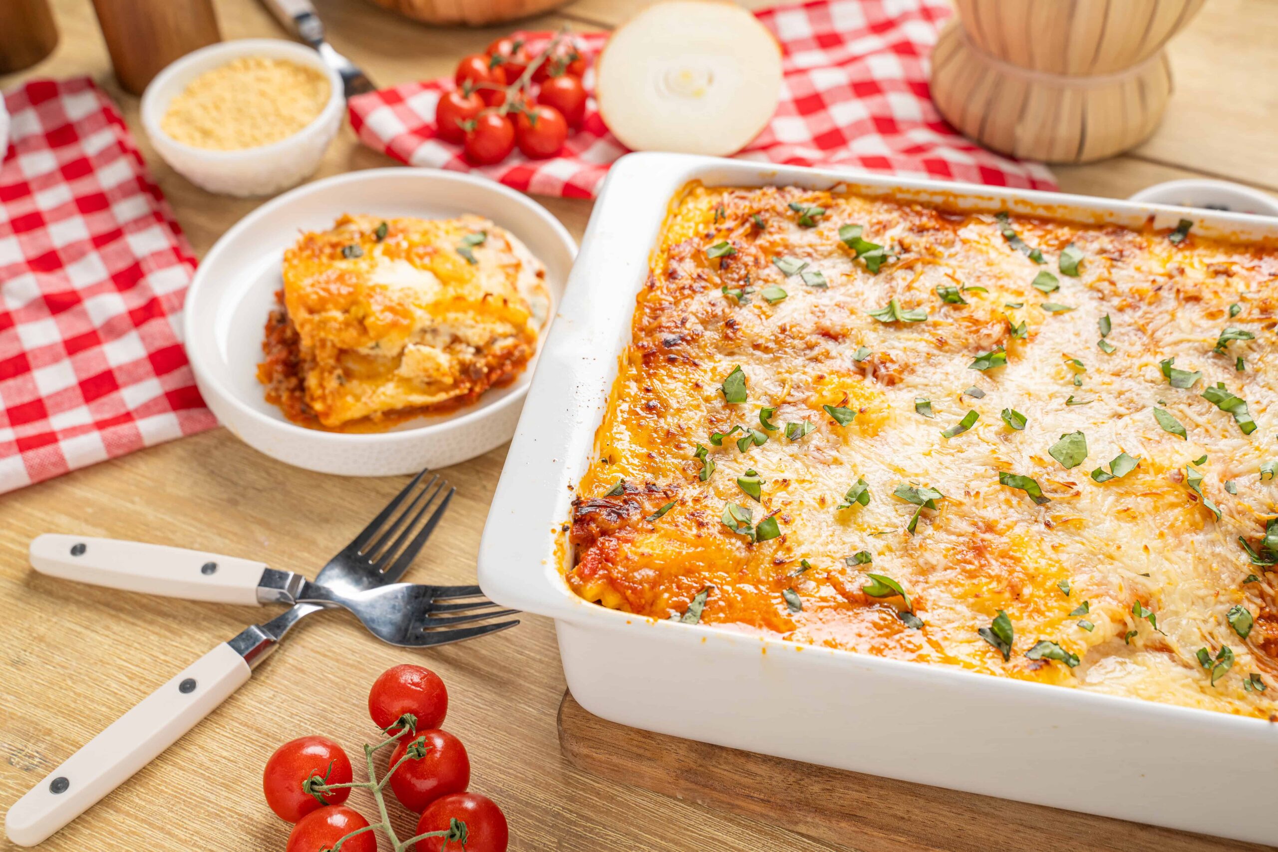 Cheesy baked lasagna with fresh herbs in a white casserole dish served with a slice on a white plate, surrounded by cherry tomatoes and cooking ingredients on a rustic wooden table.