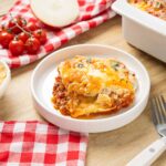Creamy lasagna with melted cheese and savory meat sauce served in a white bowl on a wooden table, surrounded by fresh ingredients and kitchen utensils, perfect for a hearty Italian meal.
