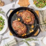 Juicy cooked steaks in a cast iron skillet served with roasted garlic, fresh rosemary, and thyme on a white marble table with green salad, olive oil, garlic, and utensils.
