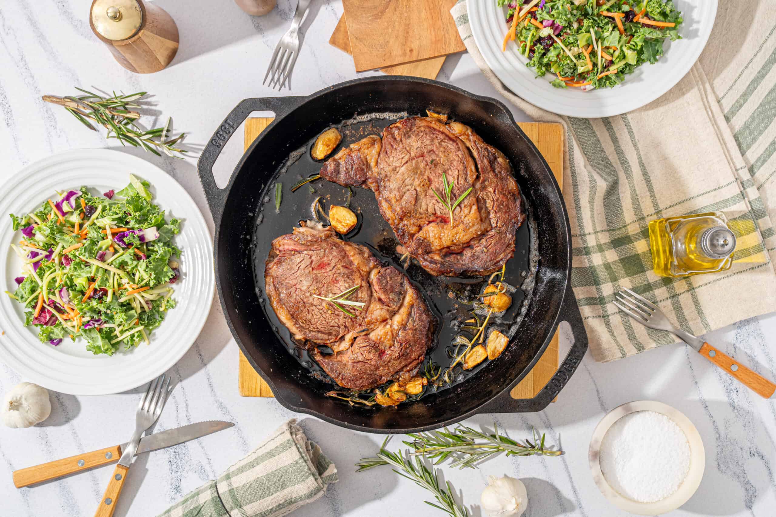 Juicy cooked steaks in a cast iron skillet served with roasted garlic, fresh rosemary, and thyme on a white marble table with green salad, olive oil, garlic, and utensils.