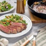 Juicy grilled steak with fresh vegetable salad on a white plate, accompanied by a cast iron skillet of beef stew and rustic table setting for a delicious home-cooked meal.