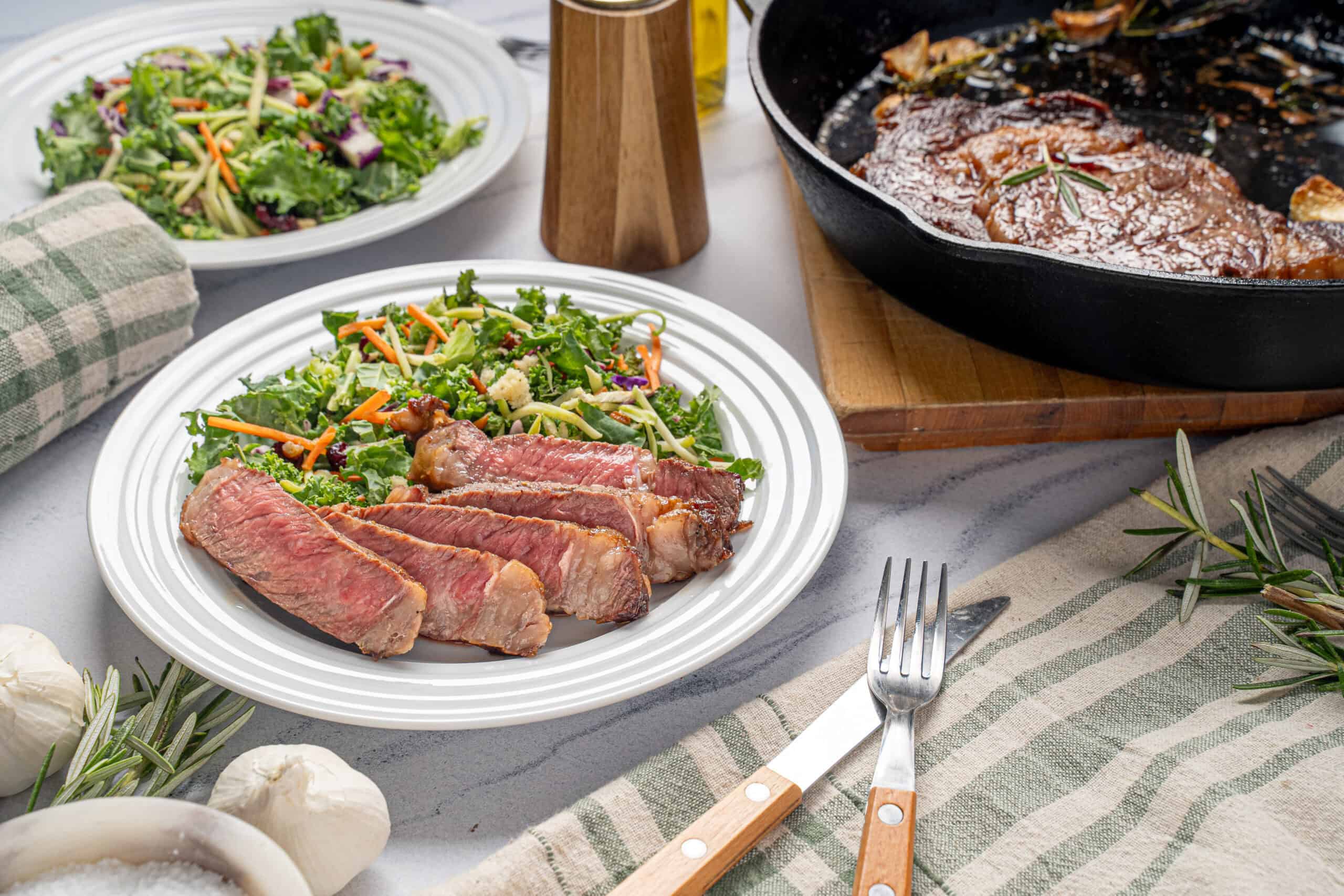 Juicy grilled steak with fresh vegetable salad on a white plate, accompanied by a cast iron skillet of beef stew and rustic table setting for a delicious home-cooked meal.