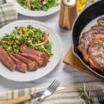 Juicy grilled steak with fresh side salad on a white plate, surrounded by garlic, herbs, and cooking ingredients, served for a delicious and healthy meal.