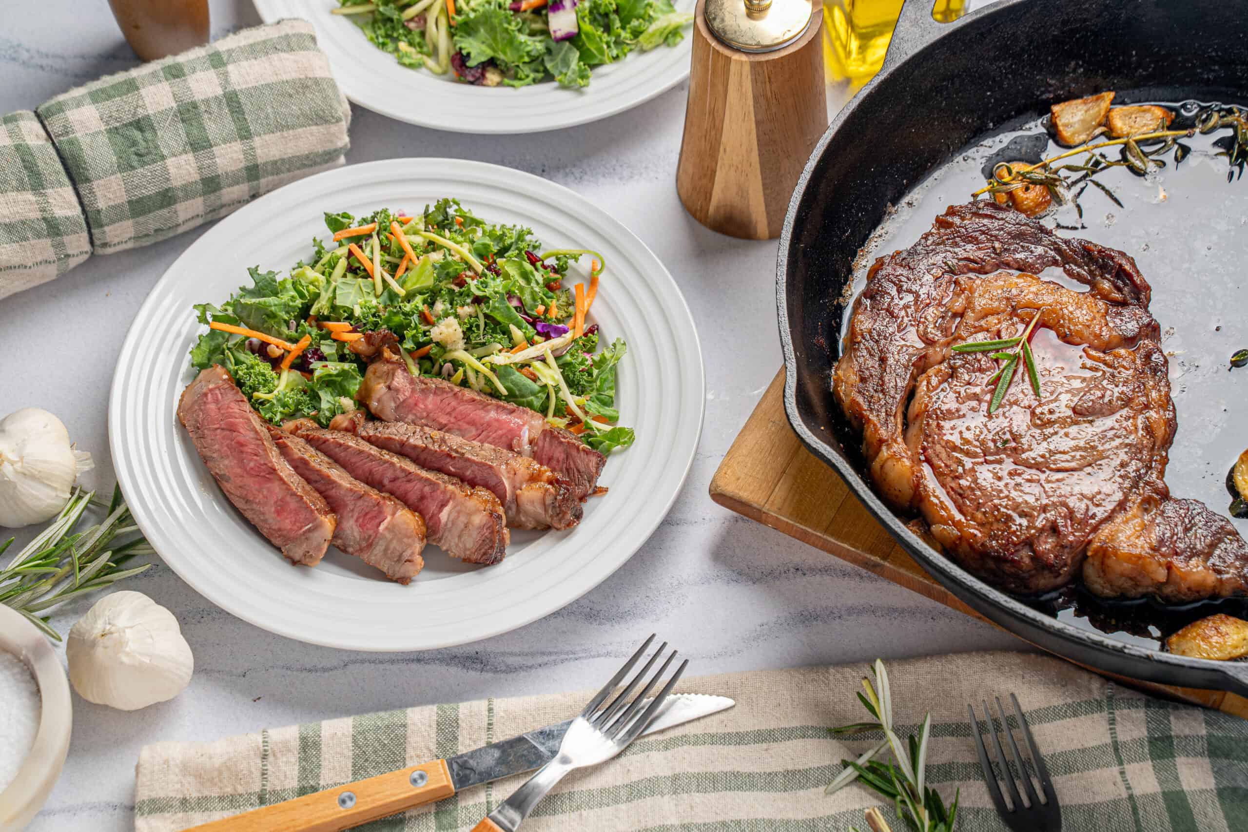 Juicy grilled steak with fresh side salad on a white plate, surrounded by garlic, herbs, and cooking ingredients, served for a delicious and healthy meal.