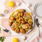 Juicy roast chicken with baby potatoes and fresh lemon slices garnished with herbs on a white plate, served with garlic and cutlery on a bright, inviting table setting.