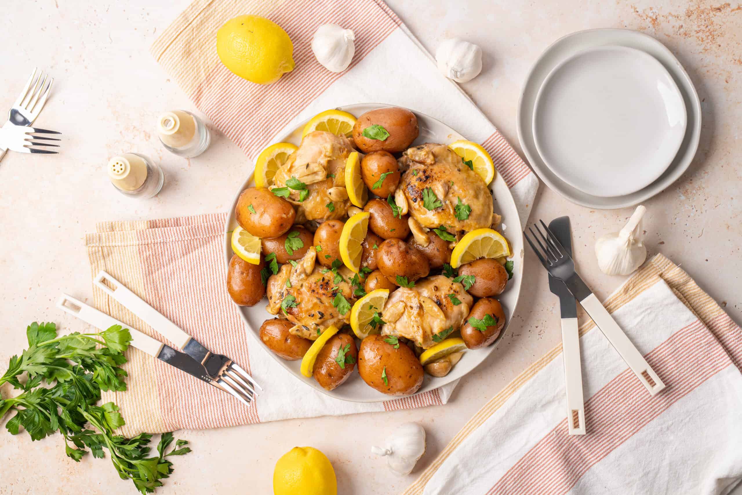 Juicy roast chicken with baby potatoes and fresh lemon slices garnished with herbs on a white plate, served with garlic and cutlery on a bright, inviting table setting.