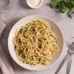 Creamy garlic spaghetti pasta topped with parsley, served on a white plate with a glass of water, salt, and fresh herbs on a textured gray surface. Perfect for quick, delicious Italian-inspired meals.